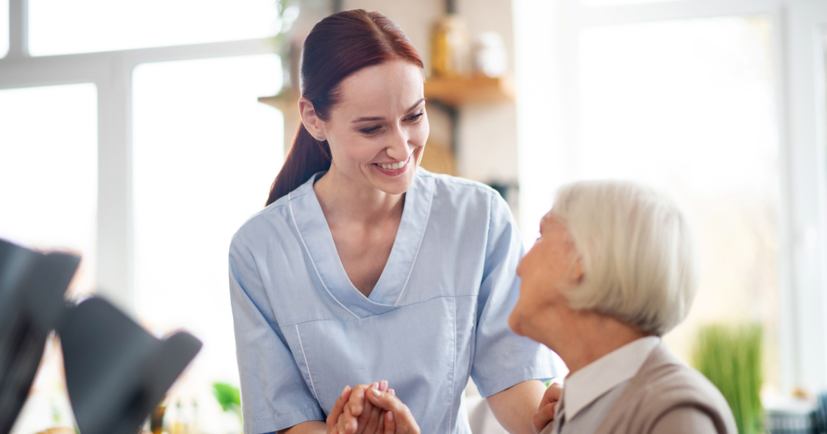 A caregiver holds hands with an elderly woman while sitting together indoors, offering comfort and support.