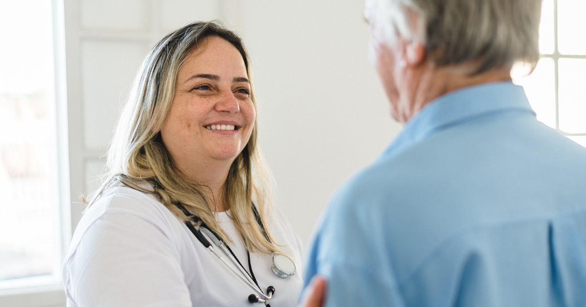 A caregiver wearing medical attire smiles while greeting an older man standing in a bright room.