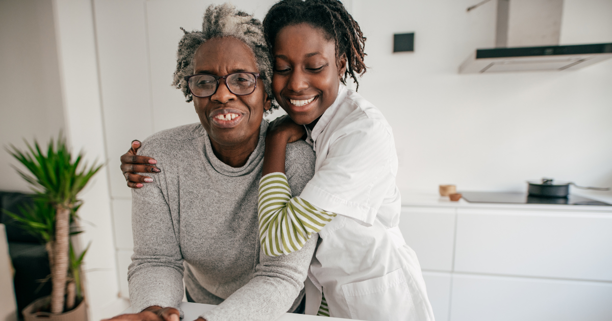 A caregiver warmly embracing a senior woman in a home kitchen, both smiling and standing close together.