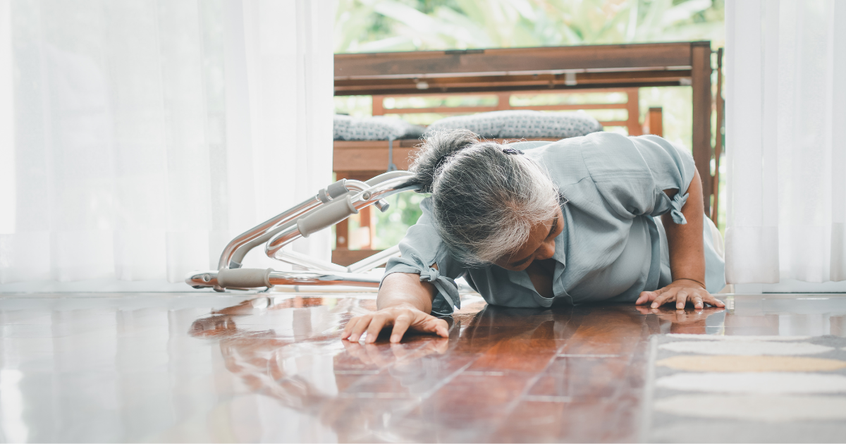 A senior woman on the floor reaching for support after a fall inside her home with natural light coming through the windows.