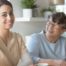 A caregiver sits at a table with an elderly woman, both holding cups of tea in a well-lit room.