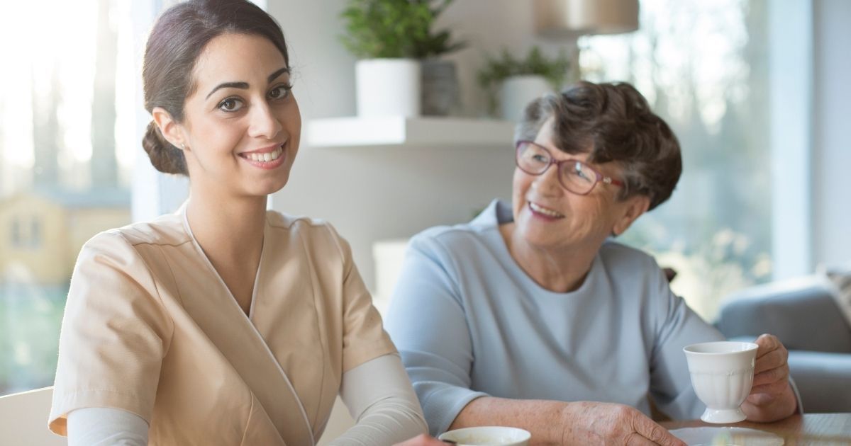 A caregiver sits at a table with an elderly woman, both holding cups of tea in a well-lit room.