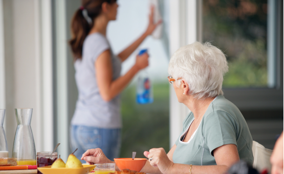 Caregiver providing light housekeeping by cleaning a window while an older woman enjoys breakfast at the table.