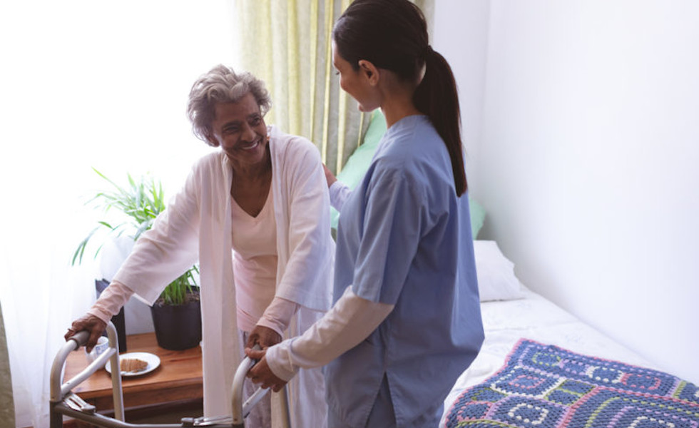 Female caregiver assisting an elderly woman as she uses a walker inside her bedroom showing personal care.