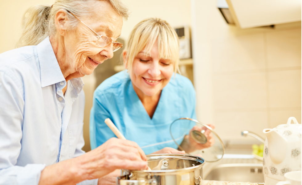 Smiling caregiver assisting an elderly woman in stirring food on the stove as part of in-home meal preparation services.