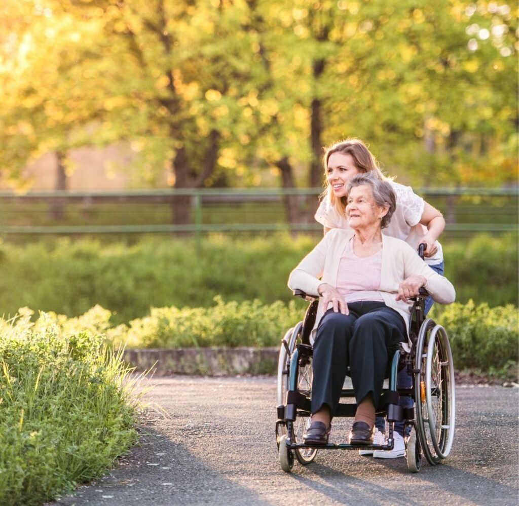 Caregiver Assisting Senior in Wheelchair During Outdoor Home Care Activity Female caregiver happily pushing an elderly woman in a wheelchair outside along a path, supporting mobility and daily home care needs.