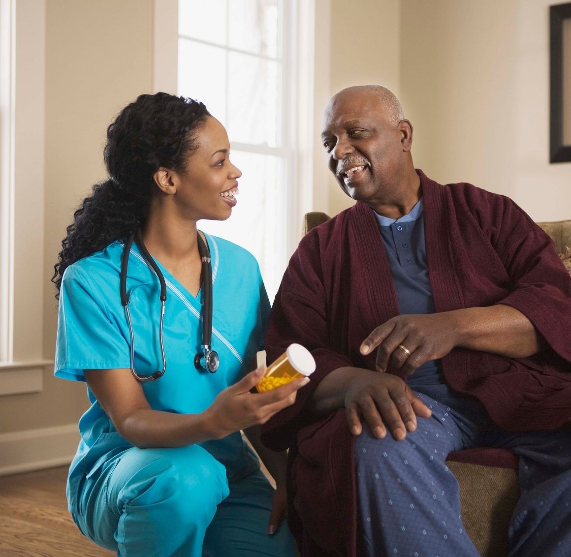 Caregiver holding a medication bottle and speaking with an elderly man while offering support at home.